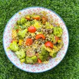 Deliciosa ensalada de pasta con brócoli, tomates cherry y semillas, servida en un cuenco con estampado geométrico sobre césped verde.