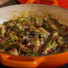 Alcachofas tiernas guisadas con trocitos de jamón, servidas en una cazuela naranja.
