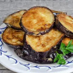 Rodajas doradas de berenjenas fritas apiladas en un plato blanco con decoración azul, adornadas con perejil fresco.