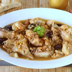 Delicioso pollo guisado con ciruelas pasas y piñones, servido en un plato blanco con pan rústico al fondo.