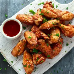 Alitas de pollo al horno crujientes, doradas y espolvoreadas con sésamo y perejil, acompañadas de una salsa roja.