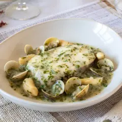 Delicioso pescado en salsa verde con almejas, servido en un plato blanco sobre un mantel de lino.
