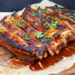 Costillas a la barbacoa jugosas y glaseadas, cubiertas con perejil fresco picado, servidas sobre papel de hornear en una tabla de madera.