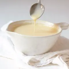 Cuchara vertiendo crema inglesa en un cuenco blanco sobre tela arrugada.
