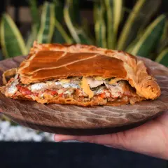 Una porción de empanada gallega, dorada y rellena de pescado y verduras, servida en plato de madera.