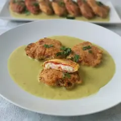 Pimientos rellenos de bacalao rebozados, servidos en una cremosa salsa verde. Uno está cortado, mostrando el relleno blanco y rojo.