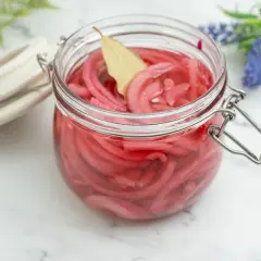 Frasco de vidrio lleno de cebolla encurtida, con una hoja de laurel flotando. Un toque de color y sabor para tus platillos.