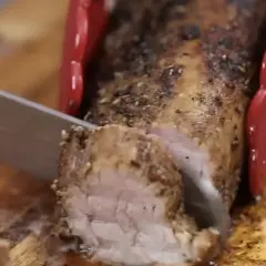 Un jugoso solomillo de cerdo al horno, recién cortado en una tabla de madera. Las pinzas rojas sujetan la carne mientras un cuchillo la rebana.