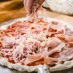 Manos esparciendo queso rallado sobre una pizza casera con jamón y salsa de tomate.