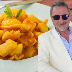 Un hombre con gafas de sol y traje blanco sonríe junto a un plato de papas con choco, un guiso amarillo con trozos de tomate y perejil.