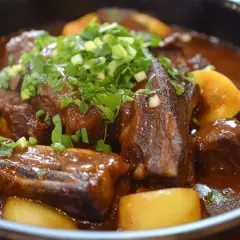 Patatas con costillas estofadas en salsa oscura, cubiertas con cebolleta picada. Un plato reconfortante y sabroso.
