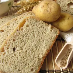 Rebanadas de pan de patata rústico, con patatas enteras, espigas de trigo y comino en una cuchara de madera.