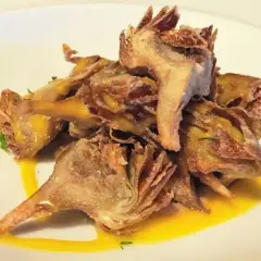 Montón de alcachofas fritas crujientes, doradas y sazonadas, servidas sobre un plato blanco con un toque de salsa amarilla y perejil picado.