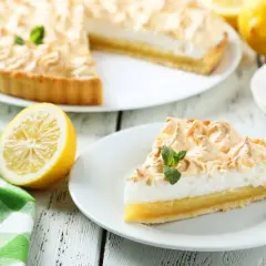 Delicioso pastel de limón con merengue dorado, decorado con menta fresca. Un trozo servido en plato blanco, junto a limones y una taza de té.