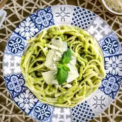 Un plato de pasta al pesto, decorado con queso parmesano y albahaca fresca, junto a una pequeña porción de salsa pesto.