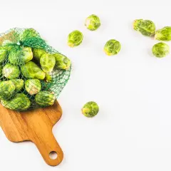 Una bolsa de malla verde llena de coles de Bruselas frescas descansa sobre una tabla de cortar de madera. Algunas coles de Bruselas están esparcidas alrededor.