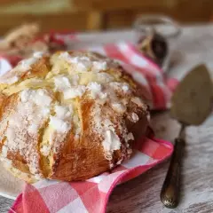 Un pan quemado dorado y crujiente, espolvoreado con azúcar, descansa sobre un plato. Una servilleta a cuadros rojos y blancos y un utensilio para pasteles completan la escena.