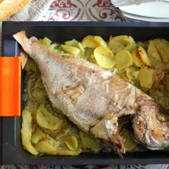 Pargo al horno con patatas y cebolla en una bandeja negra. Un pan rústico descansa a un lado.
