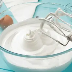 Batidora mezclando merengue italiano en un bol de cristal. Cáscara de huevo al lado.