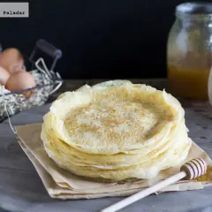 Pila de filloas gallegas recién hechas, con miel en un tarro y huevos en una cesta al fondo.