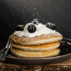 Una pila de tortitas americanas cubiertas con crema batida y moras, espolvoreadas con azúcar glas.