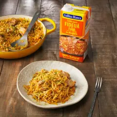 Fideos a la cazuela servidos en un plato y una paellera amarilla, junto a un caldo para fideuá.
