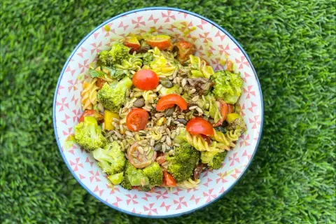Deliciosa ensalada de pasta con brócoli, tomates cherry y semillas, servida en un cuenco con estampado geométrico sobre césped verde.