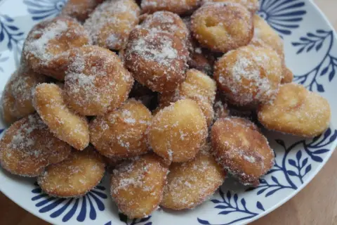Montón de buñuelos de manzana dorados y espolvoreados con azúcar, servidos en un plato blanco con diseño azul.