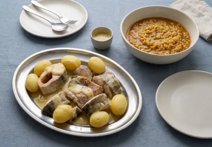 Bullit de peix servido en una bandeja de plata con patatas y una salsa cremosa, acompañado de un cuenco de arroz y un plato vacío.