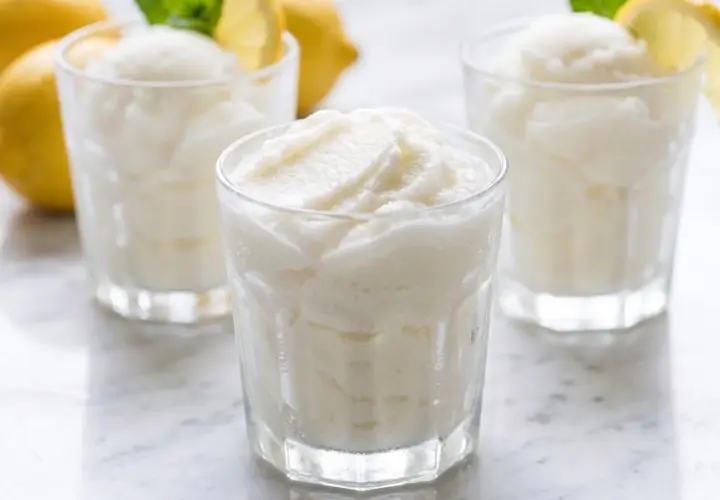 Tres vasos de sorbete de limón refrescante, con limones amarillos y hojas de menta al fondo.