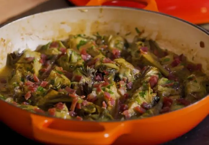Alcachofas tiernas guisadas con trocitos de jamón, servidas en una cazuela naranja.