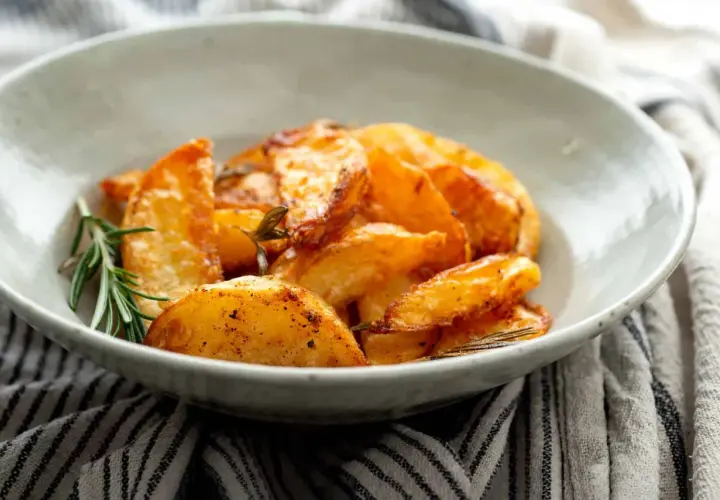 Patatas al horno en rodajas, doradas y crujientes, servidas en un cuenco con romero fresco.
