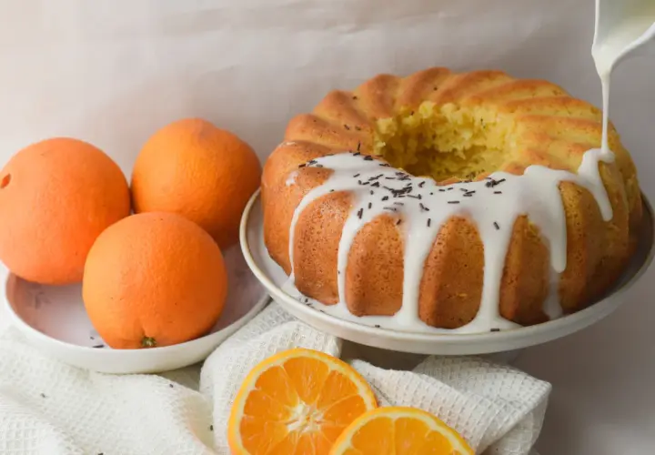 Bizcocho de naranja y yogur con glaseado blanco y chispas de chocolate. Naranjas frescas y rodajas de naranja completan la escena.