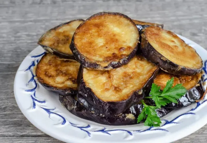 Rodajas doradas de berenjenas fritas apiladas en un plato blanco con decoración azul, adornadas con perejil fresco.