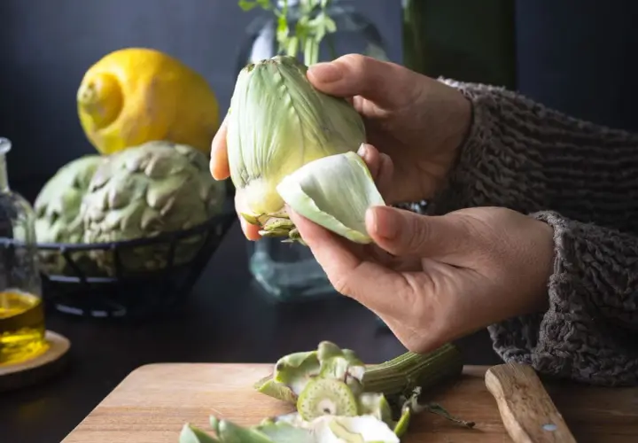 Manos preparan alcachofas para deliciosas recetas. Limón y aceite de oliva a la vista.