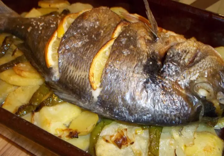 Dorada al horno con rodajas de limón, patatas y pimientos verdes en una bandeja.