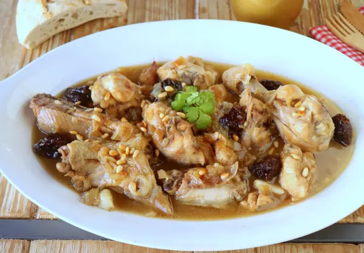 Delicioso pollo guisado con ciruelas pasas y piñones, servido en un plato blanco con pan rústico al fondo.