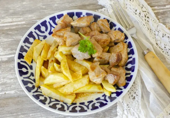 Raxo gallego jugoso con patatas fritas crujientes, servido en un plato con borde azul y blanco.
