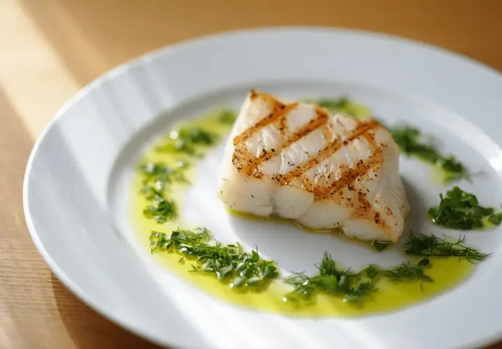 Un delicioso bacalao a la plancha, con marcas doradas, servido en un plato blanco con aceite de oliva y hierbas frescas.