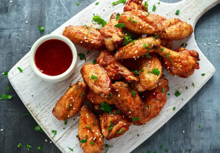 Alitas de pollo al horno crujientes, doradas y espolvoreadas con sésamo y perejil, acompañadas de una salsa roja.