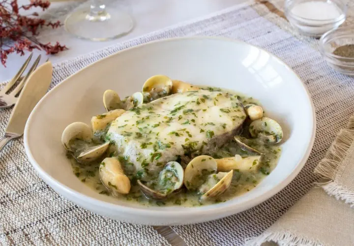 Delicioso pescado en salsa verde con almejas, servido en un plato blanco sobre un mantel de lino.