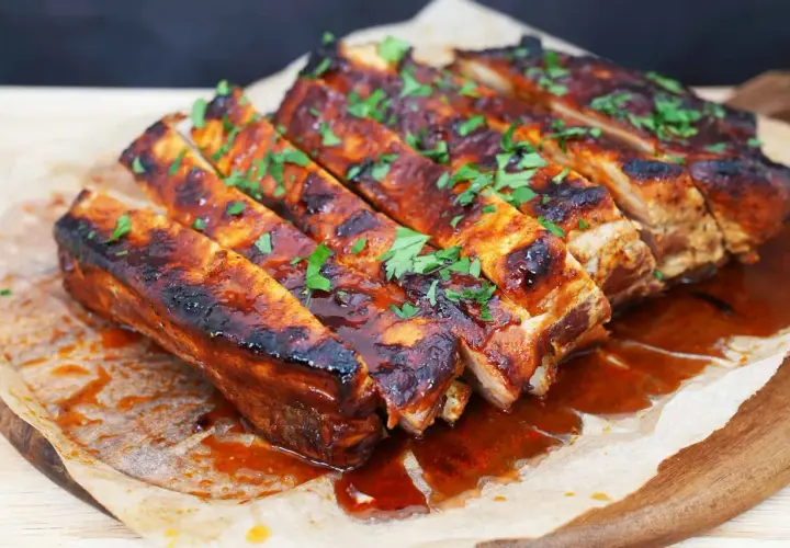 Costillas a la barbacoa jugosas y glaseadas, cubiertas con perejil fresco picado, servidas sobre papel de hornear en una tabla de madera.