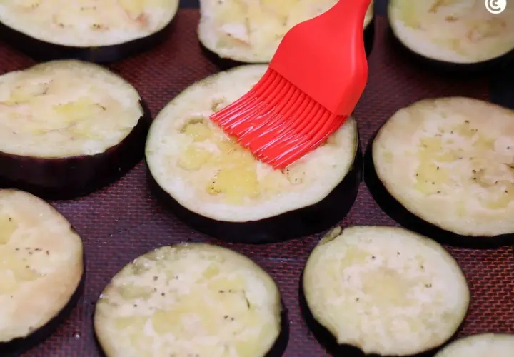 Rodajas de berenjenas listas para ser horneadas, untadas con aceite y especias con un pincel rojo.