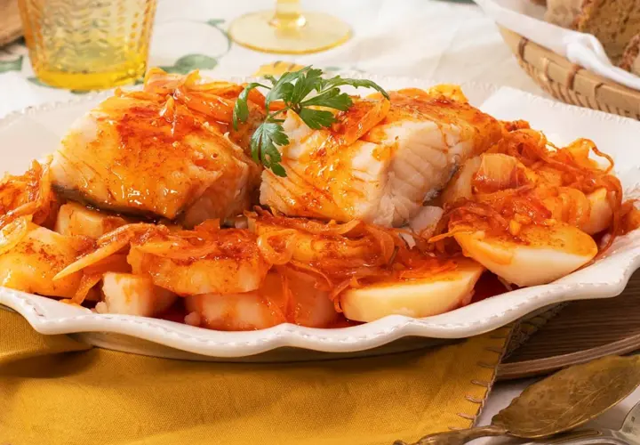 Bacalao a la gallega, un plato tradicional con trozos de pescado blanco cubiertos de una salsa rojiza con cebolla y patatas cocidas.