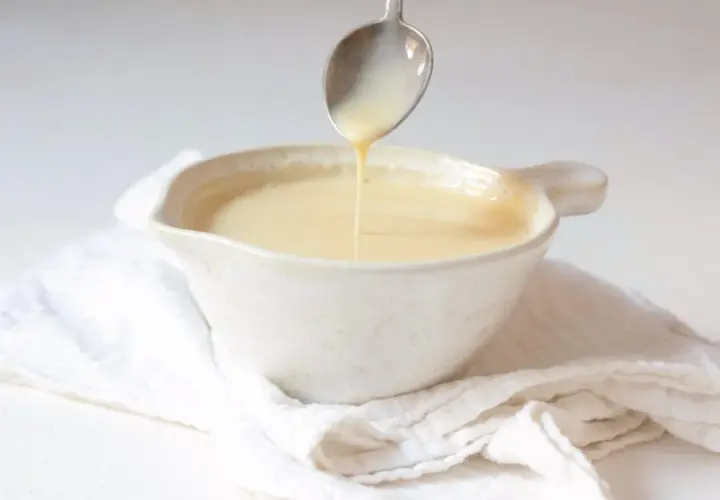 Cuchara vertiendo crema inglesa en un cuenco blanco sobre tela arrugada.