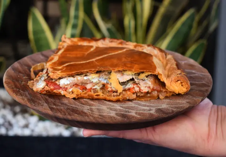 Una porción de empanada gallega, dorada y rellena de pescado y verduras, servida en plato de madera.