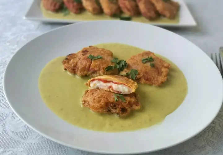 Pimientos rellenos de bacalao rebozados, servidos en una cremosa salsa verde. Uno está cortado, mostrando el relleno blanco y rojo.