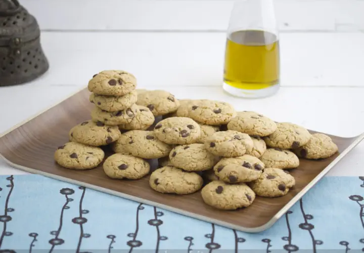 Montón de galletas caseras de pueblo con chispas de chocolate en una bandeja de madera, junto a una botella de aceite de oliva.