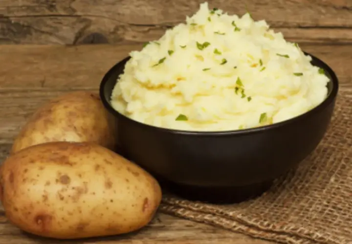 Un cremoso puré de patatas Thermomix, adornado con perejil fresco, junto a patatas crudas sobre una mesa de madera.