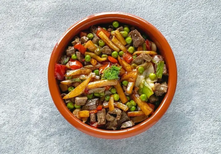 Un sabroso frito mallorquín con trozos de carne, patatas, guisantes y pimientos en una cazuela de barro.